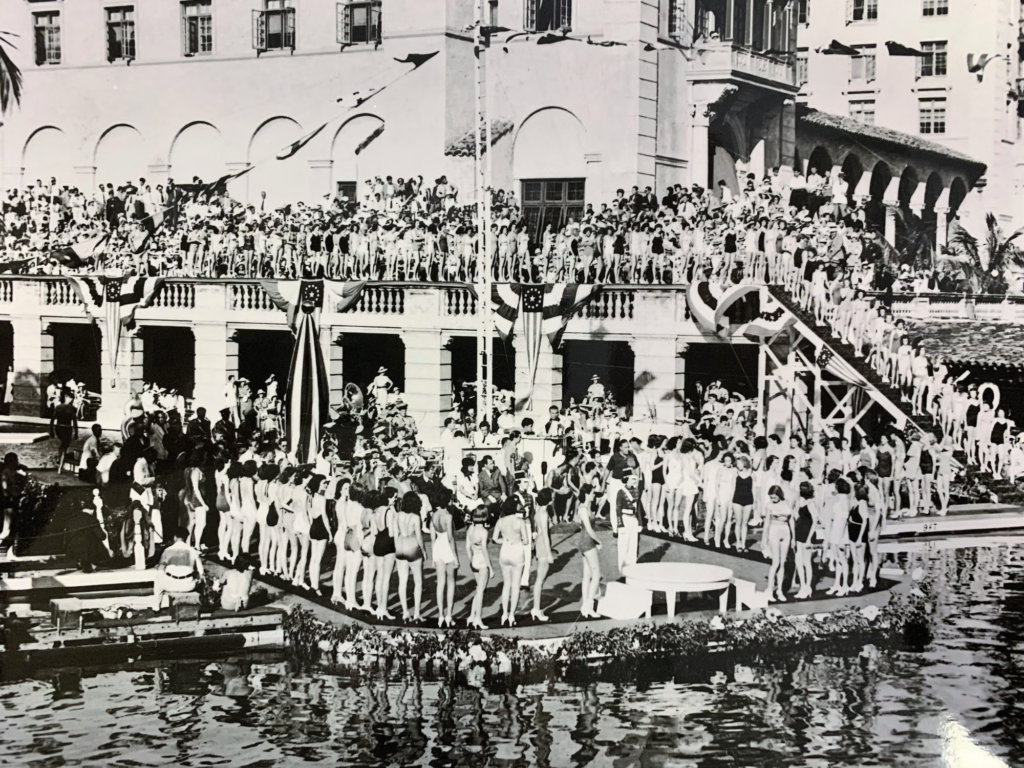 A packed aquatic spectacle transforms the Biltmore pool into a Jazz Age arena, with swimmers, spectators, patriotic bunting and a towering diving platform. The crowded historic scene offers another clue for comparing the arcade before later restorations.